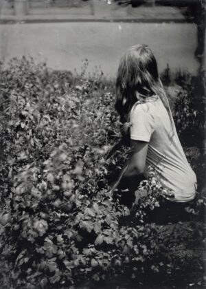 A woman in white shirt sitting on ground next to bushes.
