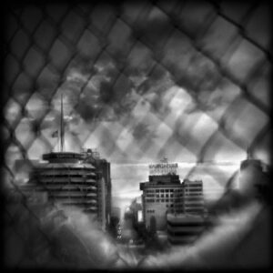 A black and white photo of buildings through a fence.
