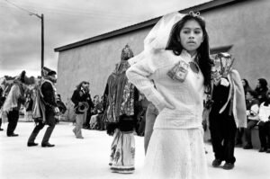 A young girl in white dress and veil standing next to crowd.
