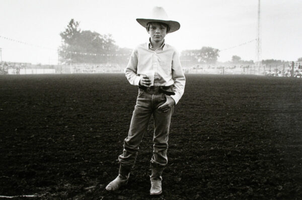 A young cowboy stands in the middle of an open field.