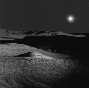 A black and white photo of the moon over some sand dunes.
