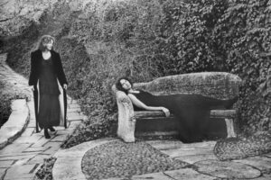 A woman in black dress laying on bench next to another person.