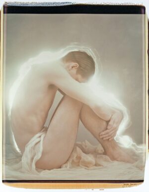 A naked man sitting on the ground with his hands in his legs.