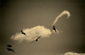 A group of birds flying in the sky with smoke coming out.