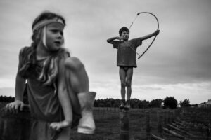 A young boy holding a bow and arrow in front of an older girl.