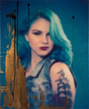 A woman with blue hair and tattoos is standing in front of a wall.