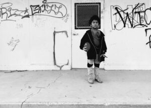 A young boy standing on the sidewalk in front of graffiti.