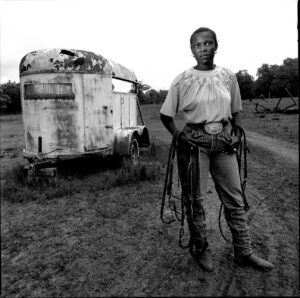 A woman standing in front of an old horse trailer.