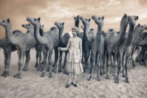 A woman standing in front of a herd of camel.