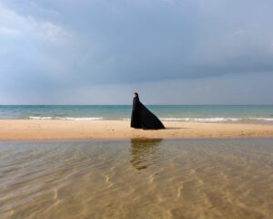 A person in black cape standing on the beach.