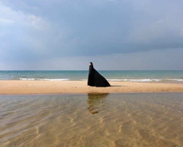 A person in black cape standing on the beach.