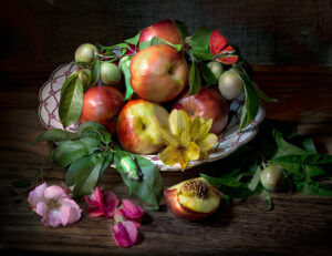 A bowl of fruit with flowers and leaves on the table.