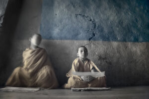 Two young monks sitting in a room with a wall behind them.
