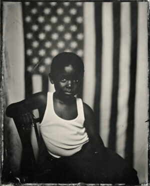 A black and white photo of a boy in front of an american flag.