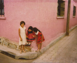 Two girls in a pink wall and one girl is bending over to look at the camera.