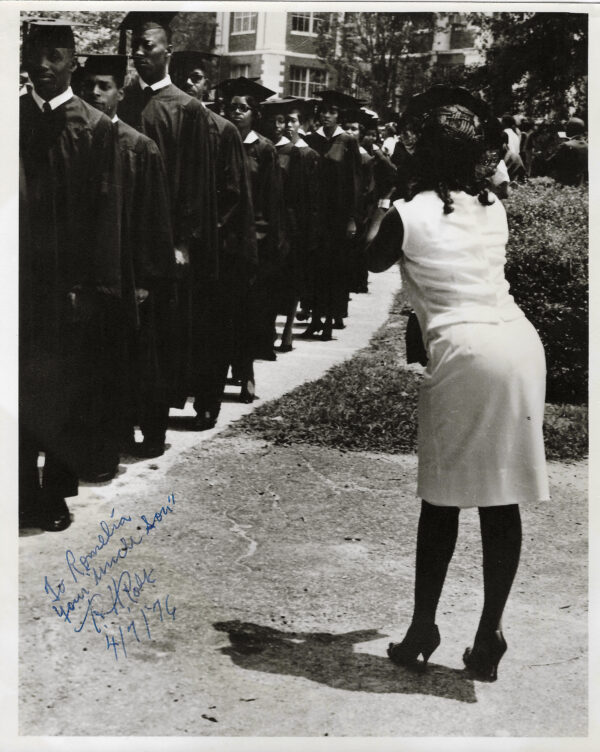 A woman in white dress standing next to a line of men.