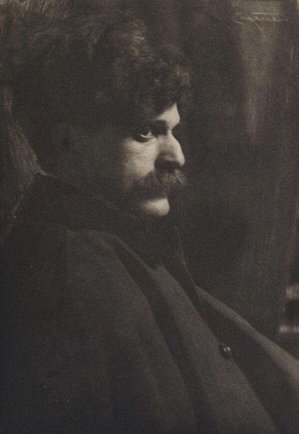 A man with a mustache sitting in front of a wall.