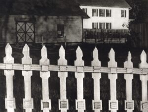 A black and white photo of a fence with houses in the background.