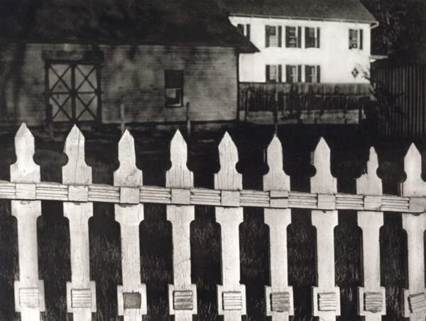 A black and white photo of a fence with houses in the background.