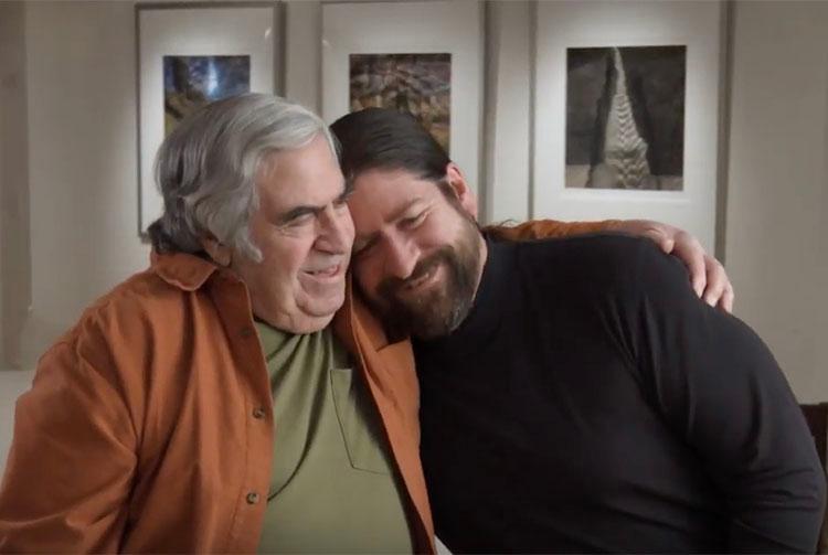 A woman and man sitting on the ground in front of a bench.