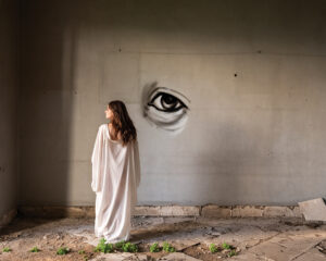 A woman in white dress standing next to wall with eye drawn on it.